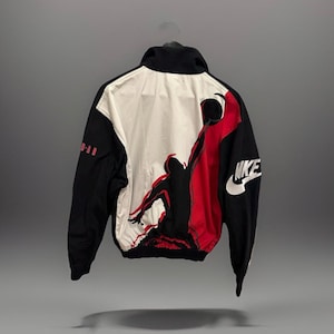 May include: Black, white, and red Jordan jacket with a silhouette graphic of a basketball player. The jacket features the Nike logo and the words "Jordan" on the sleeve. The jacket is hanging on a hanger.