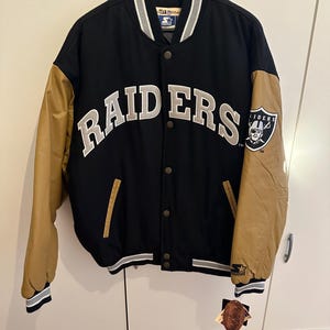 Könnte beinhalten: Schwarze und beige Raiders Collegejacke mit dem Wort "RAIDERS" in weißen und grauen Buchstaben auf der Brust. Die Jacke hat beige Ärmel, einen schwarzen Körper und einen Raiders-Logo-Aufnäher auf dem Ärmel.