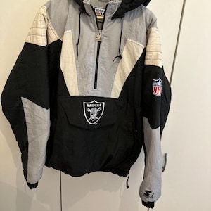 Könnte beinhalten: Eine schwarz-grau-weiße Las Vegas Raiders NFL Schlupfjacke von Starter. Dieser Kapuzen-Windbreaker verfügt über einen halben Reißverschluss, eine große Fronttasche mit Raiders-Logo, einen NFL-Aufnäher am Ärmel und ein Starter-Logo am Ärmelbündchen.