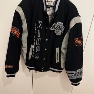 Könnte beinhalten: Schwarz-graue Los Angeles Kings Collegejacke. Die Jacke zeigt den Namen und das Logo des Teams auf der Vorderseite und den Ärmeln, mit zusätzlichen Aufnähern. Sie hat eine Knopfleiste und einen gerippten Kragen und Bündchen. Die Jacke hängt an einem schwarzen Kleiderbügel.