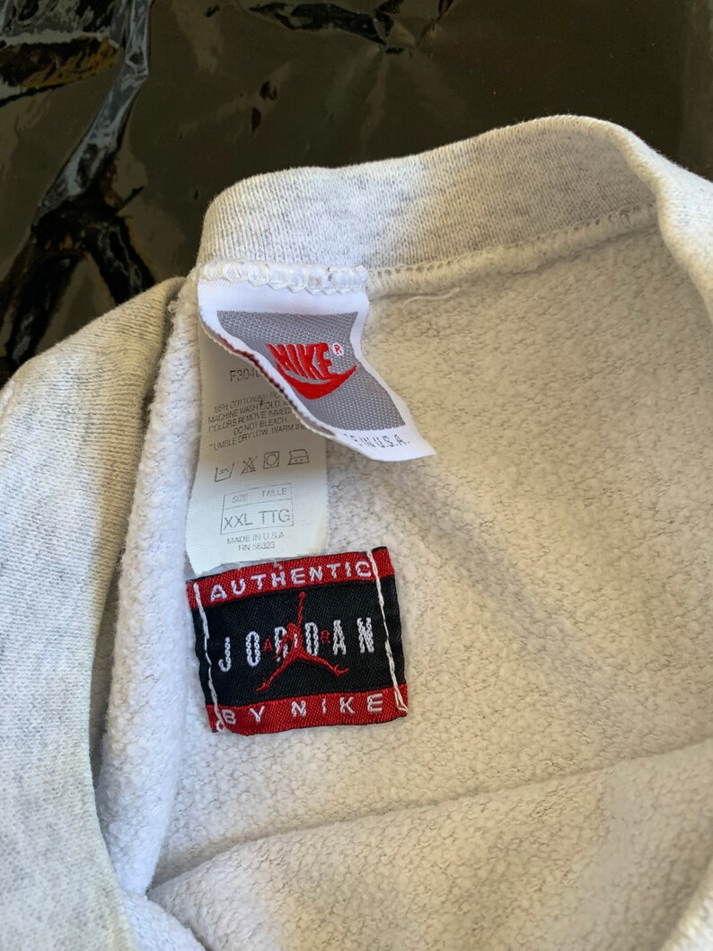 K&ouml;nnte beinhalten: Grauer Sweatshirt mit einem wei&szlig;en Etikett mit der Aufschrift "Authentic Jordan by Nike" und einem wei&szlig;en Etikett mit dem Nike-Logo und Pflegehinweisen. Das Etikett lautet "Made in U.S.A."