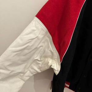 May include: A black, red, and white varsity jacket. The jacket features a red upper back and sleeves, a white leather sleeve, and a black body. The jacket has a button-up front and red and white striped trim.
