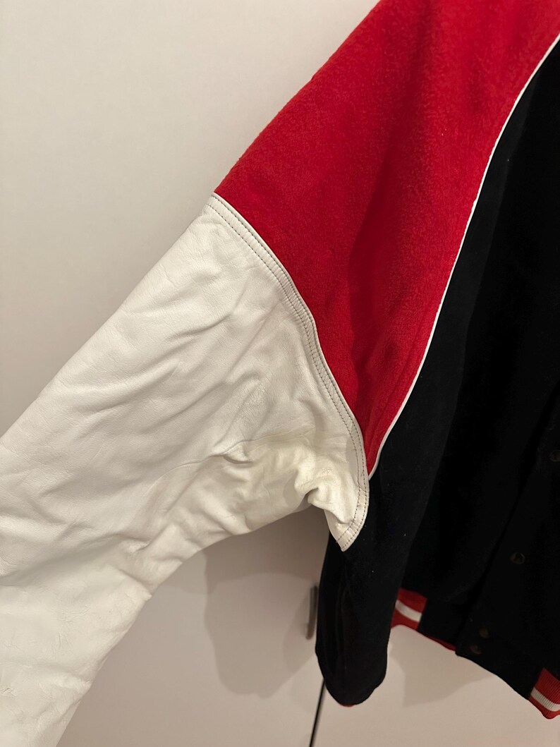 May include: A black, red, and white varsity jacket. The jacket features a red upper back and sleeves, a white leather sleeve, and a black body. The jacket has a button-up front and red and white striped trim.