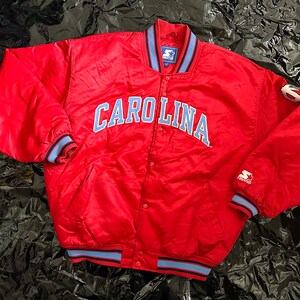 May include: Red satin bomber jacket with blue and white trim. The jacket has "CAROLINA" embroidered on the front in blue.
