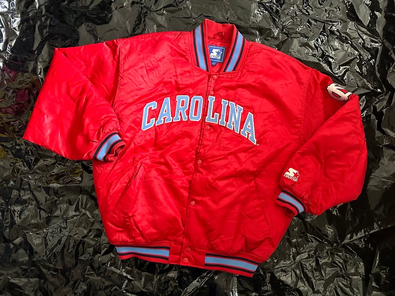 May include: Red satin bomber jacket with blue and white trim. The jacket has "CAROLINA" embroidered on the front in blue.