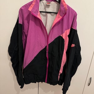 Könnte beinhalten: Vintage Nike Windjacke im Colorblock-Design. Die Jacke hat eine Kombination aus schwarzen, rosa und lila Einsätzen mit einem durchgehenden Reißverschluss. Das Nike-Logo ist auf dem Ärmel sichtbar. Die Jacke hängt an einem schwarzen Kleiderbügel.