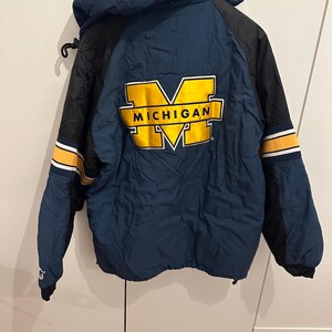 May include: A navy blue and black jacket with a hood. The jacket features the word "MICHIGAN" in yellow block letters. The sleeves have yellow and white stripes. The jacket is hanging on a black hook.