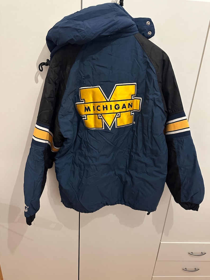 May include: A navy blue and black jacket with a hood. The jacket features the word "MICHIGAN" in yellow block letters. The sleeves have yellow and white stripes. The jacket is hanging on a black hook.