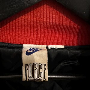 May include: Close-up of a vintage Nike jacket tag. The tag is rectangular with the word "FORCE" in bold, outlined letters. The Nike logo is above the word "FORCE". The jacket has a red ribbed collar and a black interior.