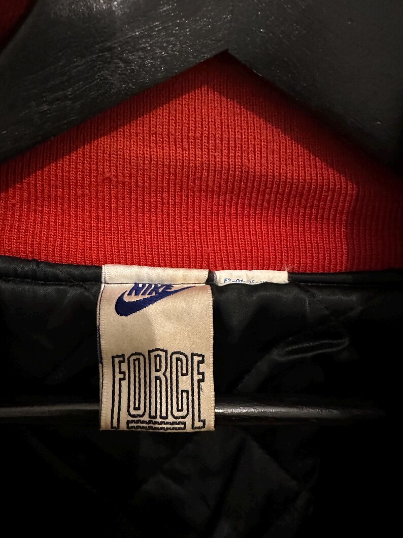 May include: Close-up of a vintage Nike jacket tag. The tag is rectangular with the word "FORCE" in bold, outlined letters. The Nike logo is above the word "FORCE". The jacket has a red ribbed collar and a black interior.