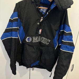 Könnte beinhalten: Schwarze und blaue Orlando Magic Starter Jacke mit Kapuze. Die Jacke zeigt das Teamlogo und das Wort "MAGIC" in Weiß. Die Ärmel haben blaue Streifen. Die Jacke hängt an einem Holzkleiderbügel.