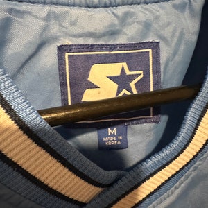 K&ouml;nnte beinhalten: Eine hellblaue Jacke mit einem wei&szlig;en und schwarzen gestreiften Kragen. Die Jacke hat einen Star-Logo-Patch und ein Etikett mit der Aufschrift "M Made in Korea".