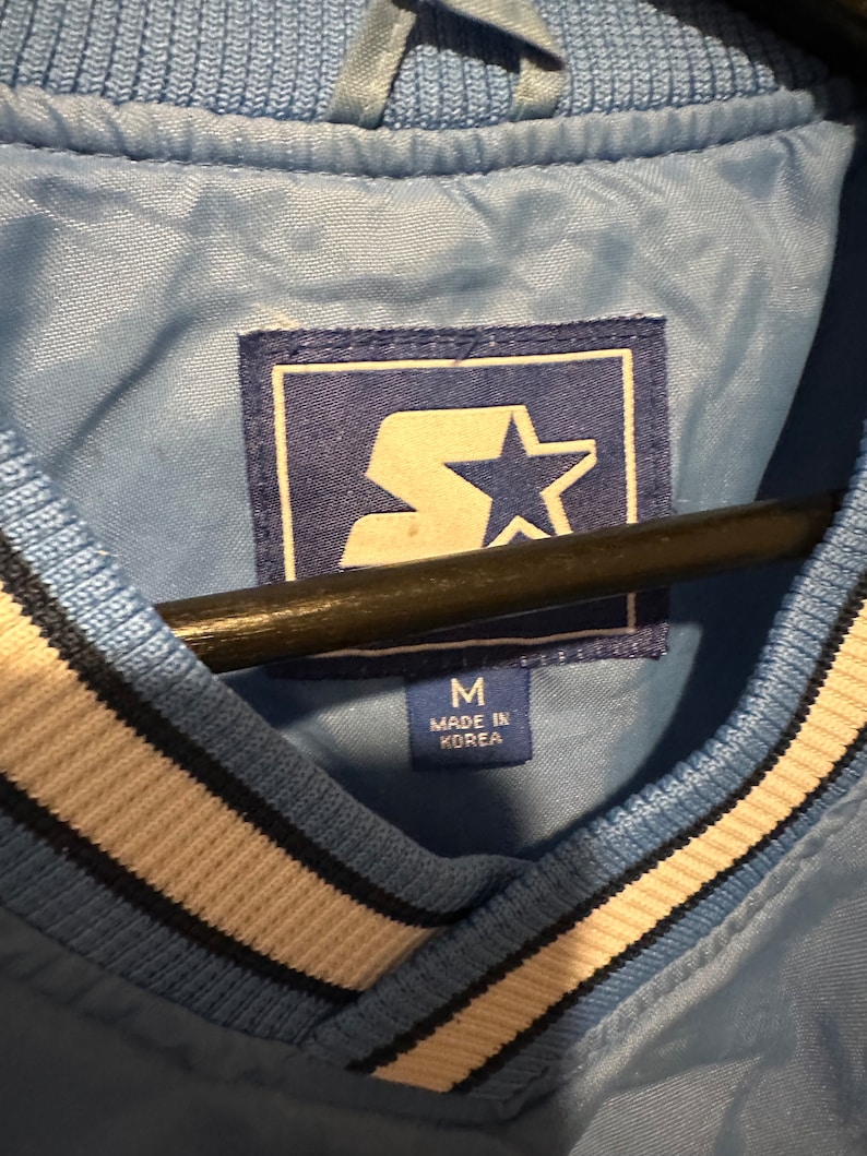 K&ouml;nnte beinhalten: Eine hellblaue Jacke mit einem wei&szlig;en und schwarzen gestreiften Kragen. Die Jacke hat einen Star-Logo-Patch und ein Etikett mit der Aufschrift "M Made in Korea".