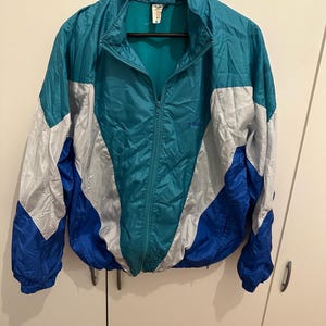 Könnte beinhalten: Eine Vintage-Trainingsjacke in Türkis, Blau und Silber mit durchgehendem Reißverschluss. Die Jacke hat ein Colorblock-Design, einen hohen Kragen und elastische Bündchen. Das Adidas-Logo ist sichtbar.