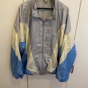 May include: A vintage Adidas track jacket in gray, light yellow, and blue. The jacket has a full zipper, a stand-up collar, and the Adidas logo in light blue on the chest. The jacket is hanging on a black hanger.