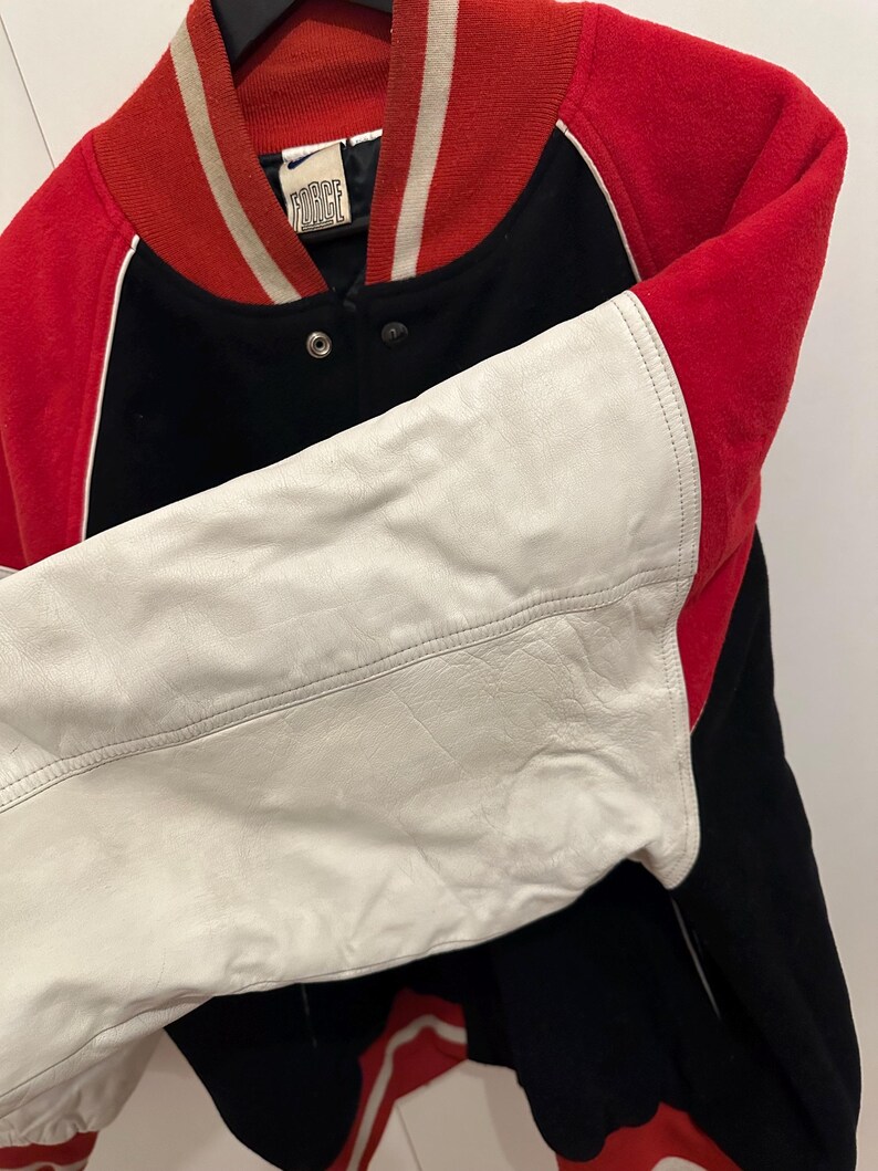 May include: A vintage letterman jacket with a red, black, and white color scheme. The jacket features a red collar and cuffs, black body, and white leather sleeves. The jacket has a snap closure and a "FORCE" label.