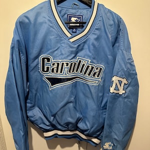 K&ouml;nnte beinhalten: Eine hellblaue Starter Windbreaker-Jacke mit einem wei&szlig;en und dunkelblauen V-Ausschnitt. Die Jacke hat ein gro&szlig;es gesticktes Carolina-Logo auf der Vorderseite und ein kleineres gesticktes UNC-Logo auf dem linken &Auml;rmel. Die Jacke hat einen wei&szlig;en und dunkelblauen gestreiften Bund und &Auml;rmelb&uuml;ndchen.