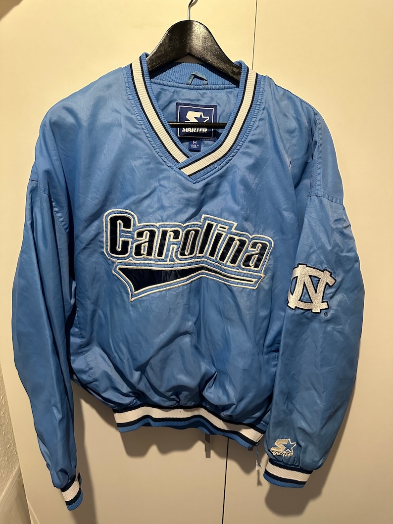 K&ouml;nnte beinhalten: Eine hellblaue Starter Windbreaker-Jacke mit einem wei&szlig;en und dunkelblauen V-Ausschnitt. Die Jacke hat ein gro&szlig;es gesticktes Carolina-Logo auf der Vorderseite und ein kleineres gesticktes UNC-Logo auf dem linken &Auml;rmel. Die Jacke hat einen wei&szlig;en und dunkelblauen gestreiften Bund und &Auml;rmelb&uuml;ndchen.