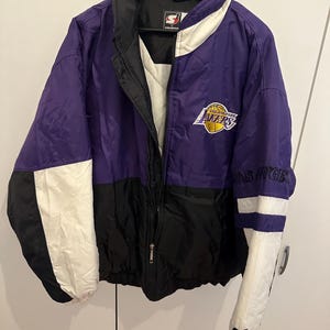 Puede incluir: Chaqueta Starter de los Lakers de Los Ángeles, morada, negra y blanca. La chaqueta presenta el logotipo de los Lakers en el pecho y la inscripción "LOS ANGELES" en la manga. Tiene cremallera completa y cuello negro.