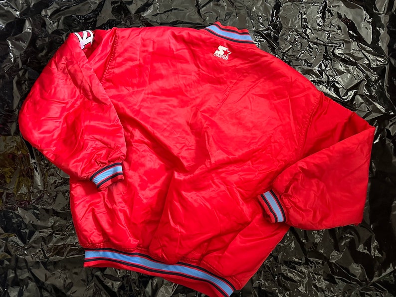 May include: A red satin bomber jacket with blue and white striped trim around the collar, cuffs, and hem. The jacket has a StarTech logo on the back.