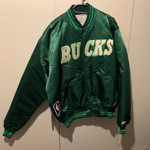 May include: Green satin bomber jacket with the word "BUCKS" in white and green lettering on the front. The jacket has a green and white striped collar and cuffs. There is a small NBA logo patch on the left sleeve.