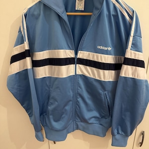 Puede incluir: Una chaqueta de chándal Adidas vintage azul claro con rayas blancas y negras y el logotipo de Adidas en el pecho.