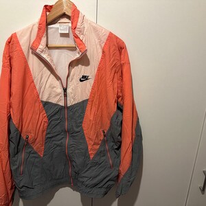 Può includere: Una giacca a vento Nike vintage in un design a blocchi di colore con pannelli rosa, arancio e grigio. La giacca ha una chiusura a zip e un logo Nike sul petto.