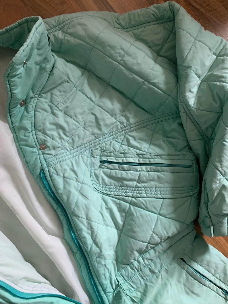 May include: A quilted mint green jacket with a zipper closure and a zippered pocket on the left side. The jacket has a high collar and a white lining.