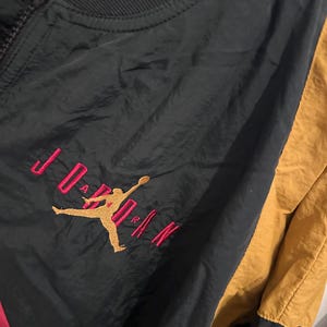 K&ouml;nnte beinhalten: Schwarze, goldene und rote Jordan-Jacke mit durchgehendem Rei&szlig;verschluss. Die Jacke hat das Jordan-Logo in Gold mit rotem Text. Die Jacke hat einen schwarzen Kragen und einen goldenen Einsatz auf der rechten Seite. Die Jacke besteht aus gl&auml;nzendem Material.