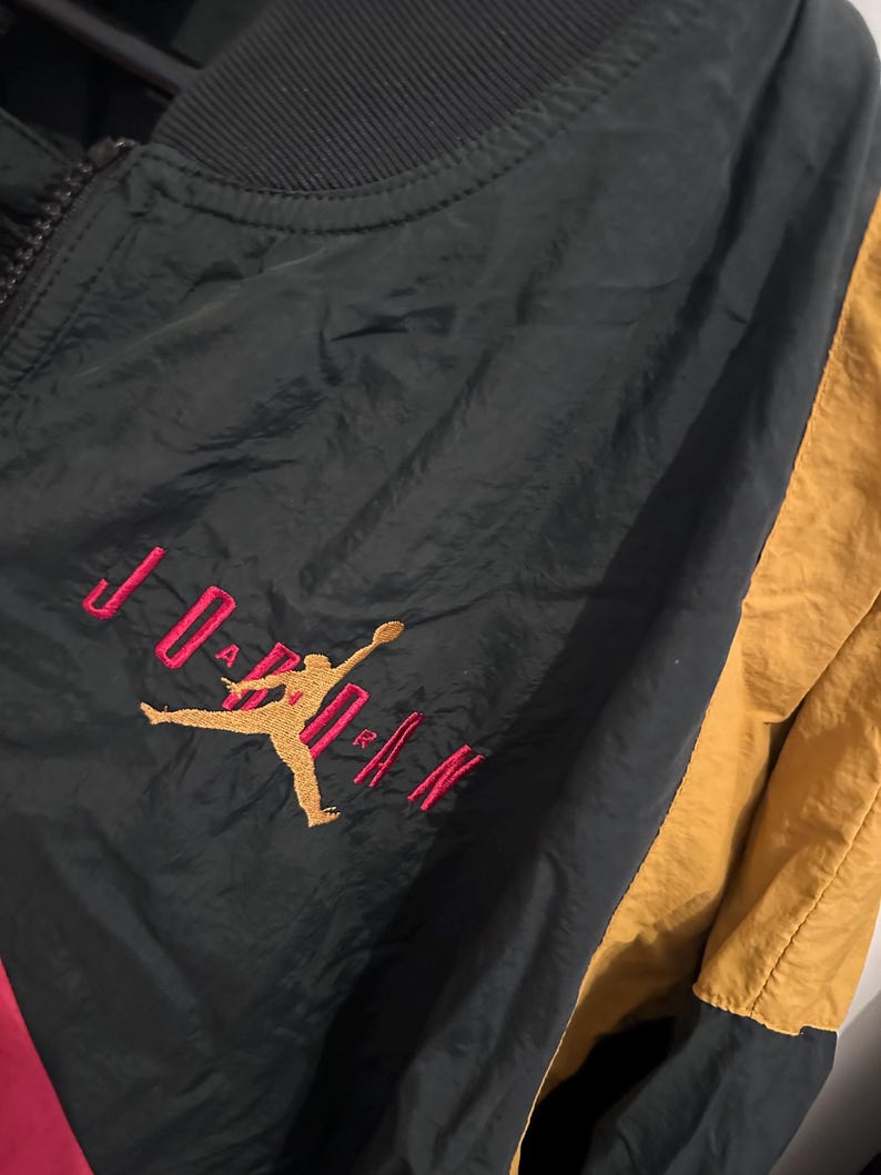 K&ouml;nnte beinhalten: Schwarze, goldene und rote Jordan-Jacke mit durchgehendem Rei&szlig;verschluss. Die Jacke hat das Jordan-Logo in Gold mit rotem Text. Die Jacke hat einen schwarzen Kragen und einen goldenen Einsatz auf der rechten Seite. Die Jacke besteht aus gl&auml;nzendem Material.
