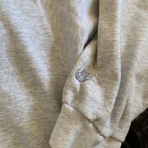 K&ouml;nnte beinhalten: Grauer Sweatshirt mit einem gestickten Nike-Logo auf dem &Auml;rmel.