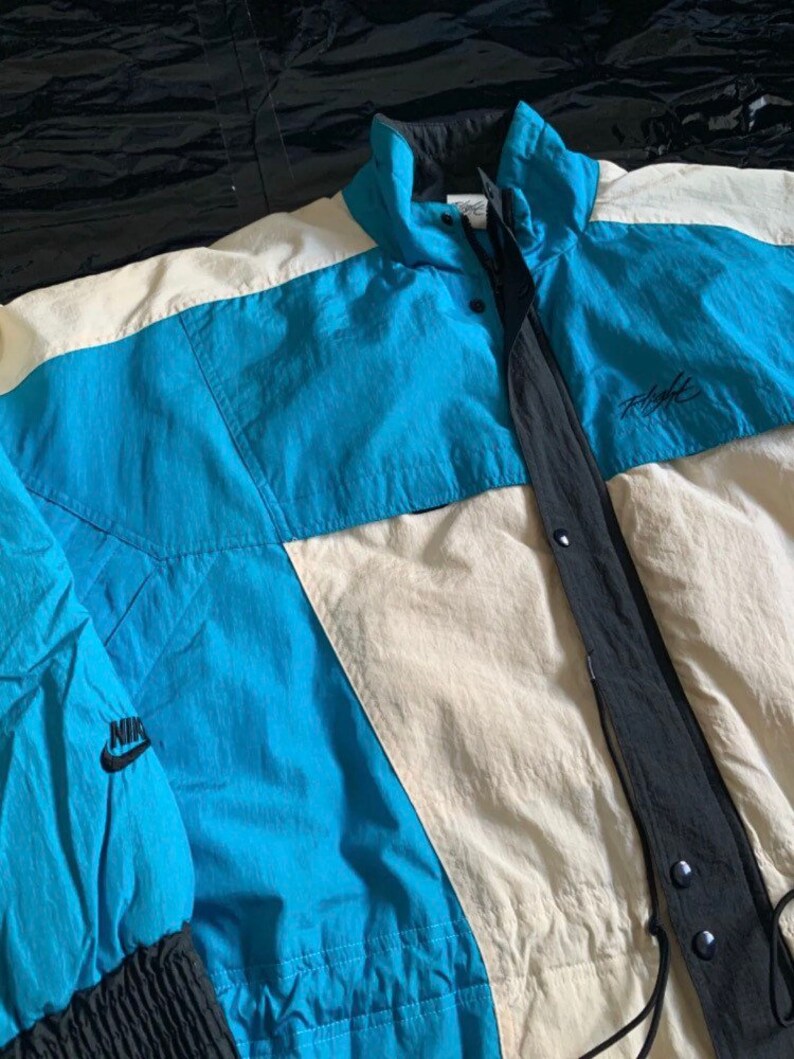May include: A vintage Nike Flight jacket in blue, white, and black with a black Nike logo on the left sleeve. The jacket has a zipper closure and a drawstring hood.