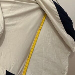 Puede incluir: Una chaqueta blanca con detalles en azul y marino. Una cinta m&eacute;trica amarilla est&aacute; colocada sobre la chaqueta. La chaqueta tiene una franja azul claro en el hombro. La chaqueta es de material ligero.