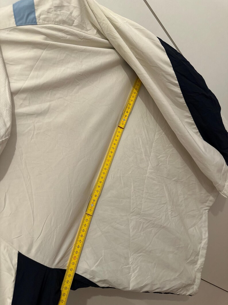 Puede incluir: Una chaqueta blanca con detalles en azul y marino. Una cinta m&eacute;trica amarilla est&aacute; colocada sobre la chaqueta. La chaqueta tiene una franja azul claro en el hombro. La chaqueta es de material ligero.