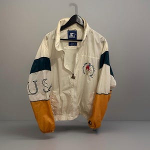Puede incluir: Chaqueta Starter vintage color crema con cremallera completa. La chaqueta presenta detalles en verde oscuro y dorado en las mangas, con "US" impreso en una manga. La otra manga tiene el logo olímpico de Atlanta 1996.