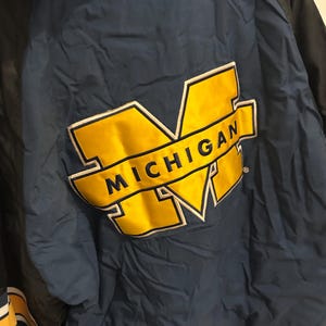 May include: A navy blue and black jacket with a large yellow and white "MICHIGAN" logo on the back. The jacket has a quilted texture and a striped yellow and white detail on the sleeve.