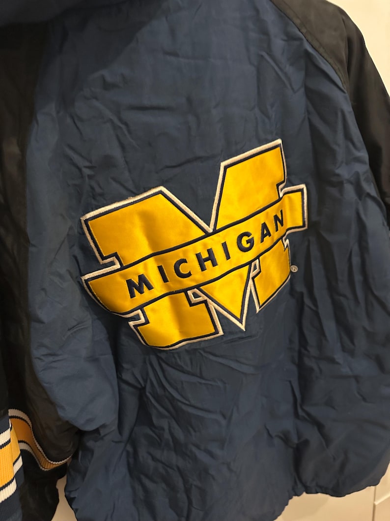 May include: A navy blue and black jacket with a large yellow and white "MICHIGAN" logo on the back. The jacket has a quilted texture and a striped yellow and white detail on the sleeve.