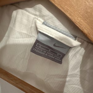 Puede incluir: Primer plano de una etiqueta de prenda blanca con el logo de Nike. La etiqueta es gris y azul, con el texto "MADE IN HONG KONG" y "FABRIQUE A HONG KONG". La prenda es blanca y parece estar colgada de una percha de madera.