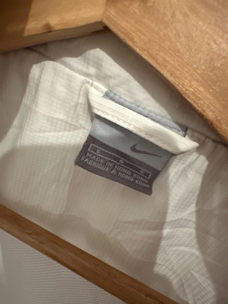 Puede incluir: Primer plano de una etiqueta de prenda blanca con el logo de Nike. La etiqueta es gris y azul, con el texto "MADE IN HONG KONG" y "FABRIQUE A HONG KONG". La prenda es blanca y parece estar colgada de una percha de madera.