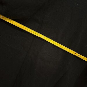 May include: A black fabric is measured with a yellow measuring tape. The tape is marked in centimetres and is laid diagonally across the fabric. The fabric appears to be a solid colour.