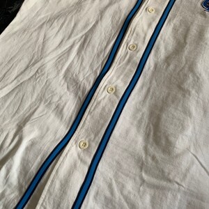 May include: White baseball jersey with blue and black stripes on the sleeves and down the front. The jersey has a button-up front and a small logo on the right side of the chest.