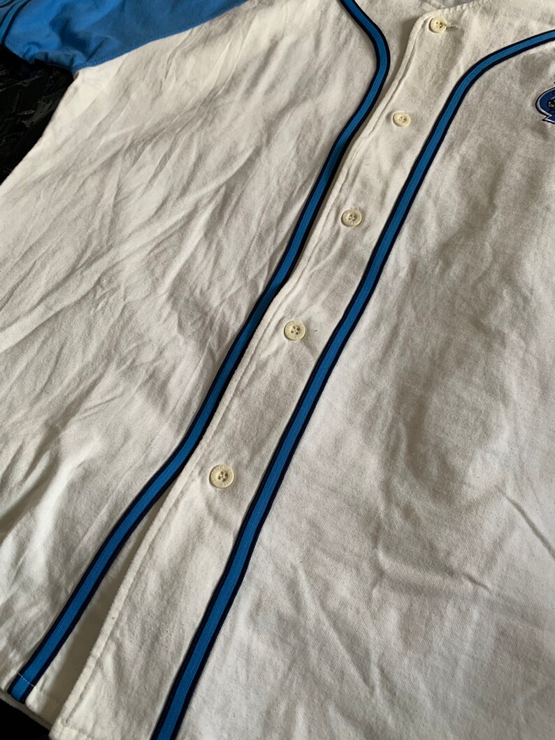 May include: White baseball jersey with blue and black stripes on the sleeves and down the front. The jersey has a button-up front and a small logo on the right side of the chest.