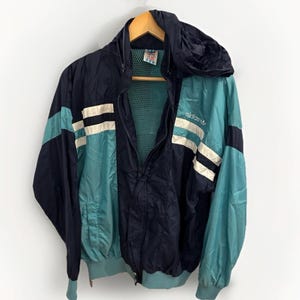 Op de afbeelding: Een blauwe en turquoise vintage Adidas windbreaker jack met witte strepen en een mesh voering. De jas heeft een capuchon en een ritssluiting. Het Adidas logo staat op de rechterkant van de borst.