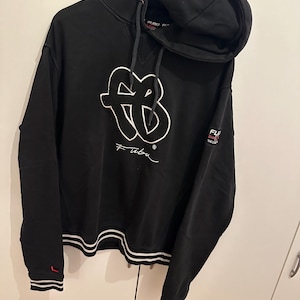 以下が含まれることがあります： 白の刺繍ロゴとブランド名「Fubu」が特徴の黒いフード付きスウェットシャツ。袖口と裾には白いストライプが入っています。左袖口には小さな赤い正方形があります。