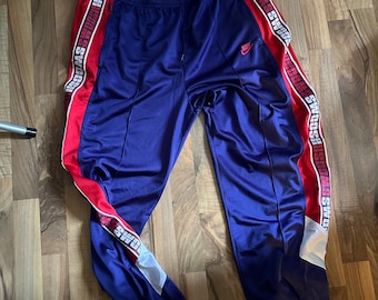 May include: Blue sweatpants with red and white stripes down the sides. The stripes have the word "SWOOSH" repeated on them. The sweatpants have a white Nike logo on the left leg.