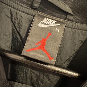 K&ouml;nnte beinhalten: Nahaufnahme eines schwarzen Jackenetiketts mit dem wei&szlig;en Nike-Logo und dem roten Jumpman-Logo. Das Etikett weist auch die Gr&ouml;&szlig;e XL aus. Die Jacke scheint aus einem gl&auml;nzenden, wasserabweisenden Material zu bestehen.