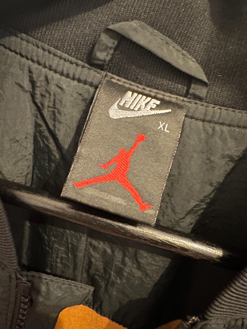 K&ouml;nnte beinhalten: Nahaufnahme eines schwarzen Jackenetiketts mit dem wei&szlig;en Nike-Logo und dem roten Jumpman-Logo. Das Etikett weist auch die Gr&ouml;&szlig;e XL aus. Die Jacke scheint aus einem gl&auml;nzenden, wasserabweisenden Material zu bestehen.
