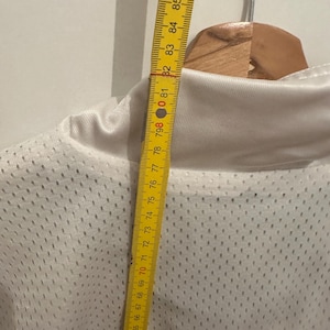 Peut inclure: Un maillot en maille blanche avec un motif rouge et noir en bas. Un m&egrave;tre ruban jaune indique que la longueur du maillot du col au bas est d'environ 76 cm.