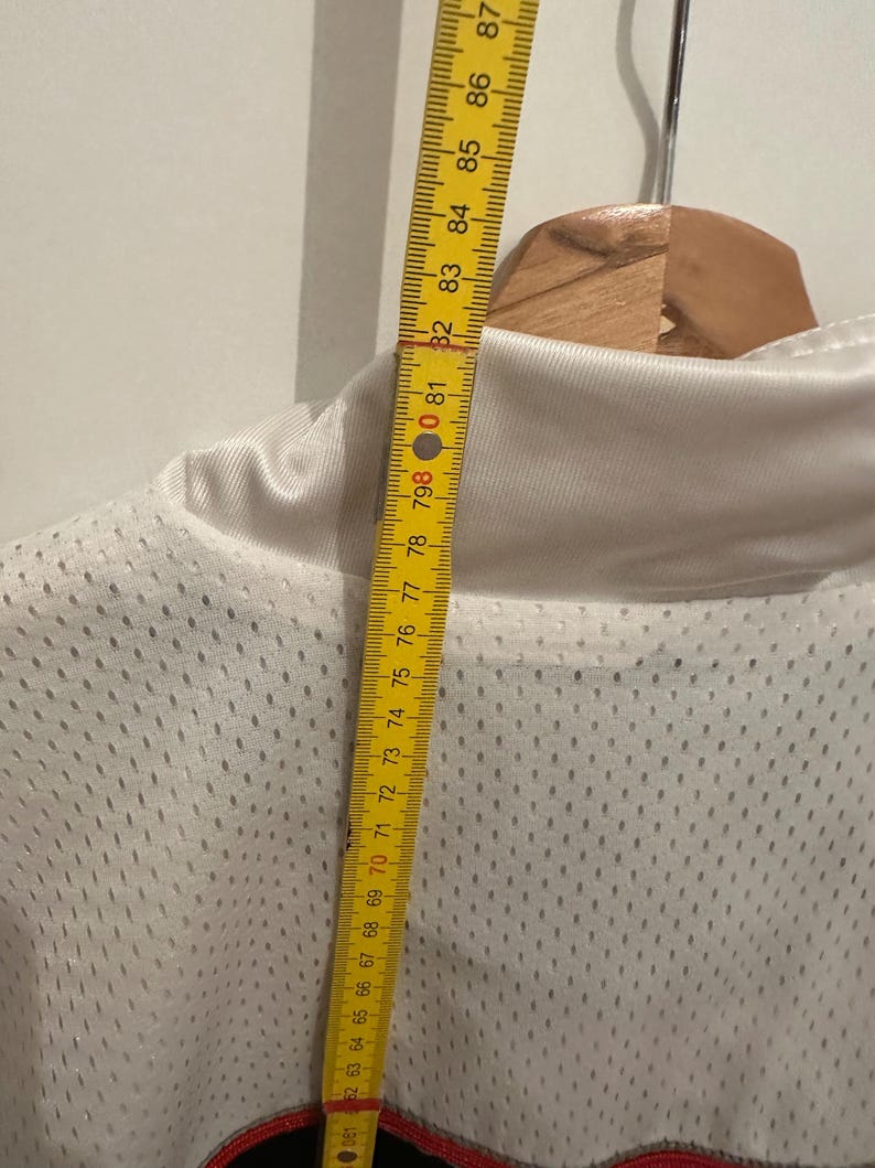 Peut inclure: Un maillot en maille blanche avec un motif rouge et noir en bas. Un m&egrave;tre ruban jaune indique que la longueur du maillot du col au bas est d'environ 76 cm.