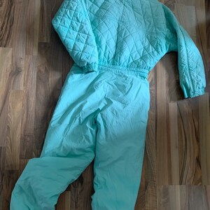 May include: A light blue quilted ski suit with a zipper closure. The suit has a quilted top and matching pants.